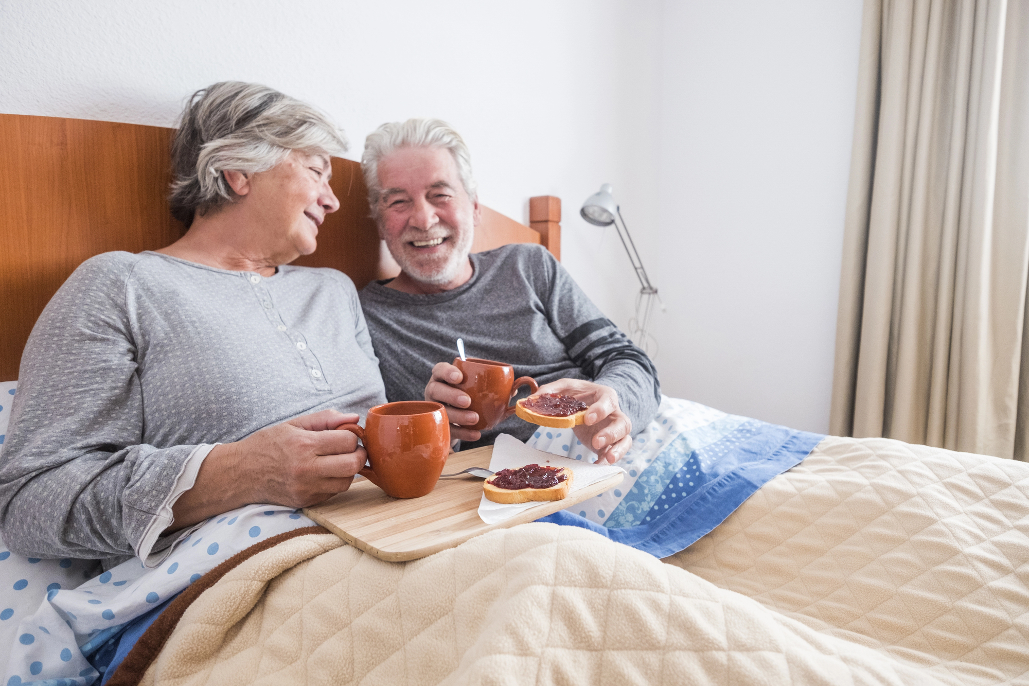 An elderly couple is sitting in bed under a beige blanket, smiling and enjoying breakfast. They have mugs and a tray with toast and jam. The bed has blue and polka dot sheets, and a lamp is in the background.