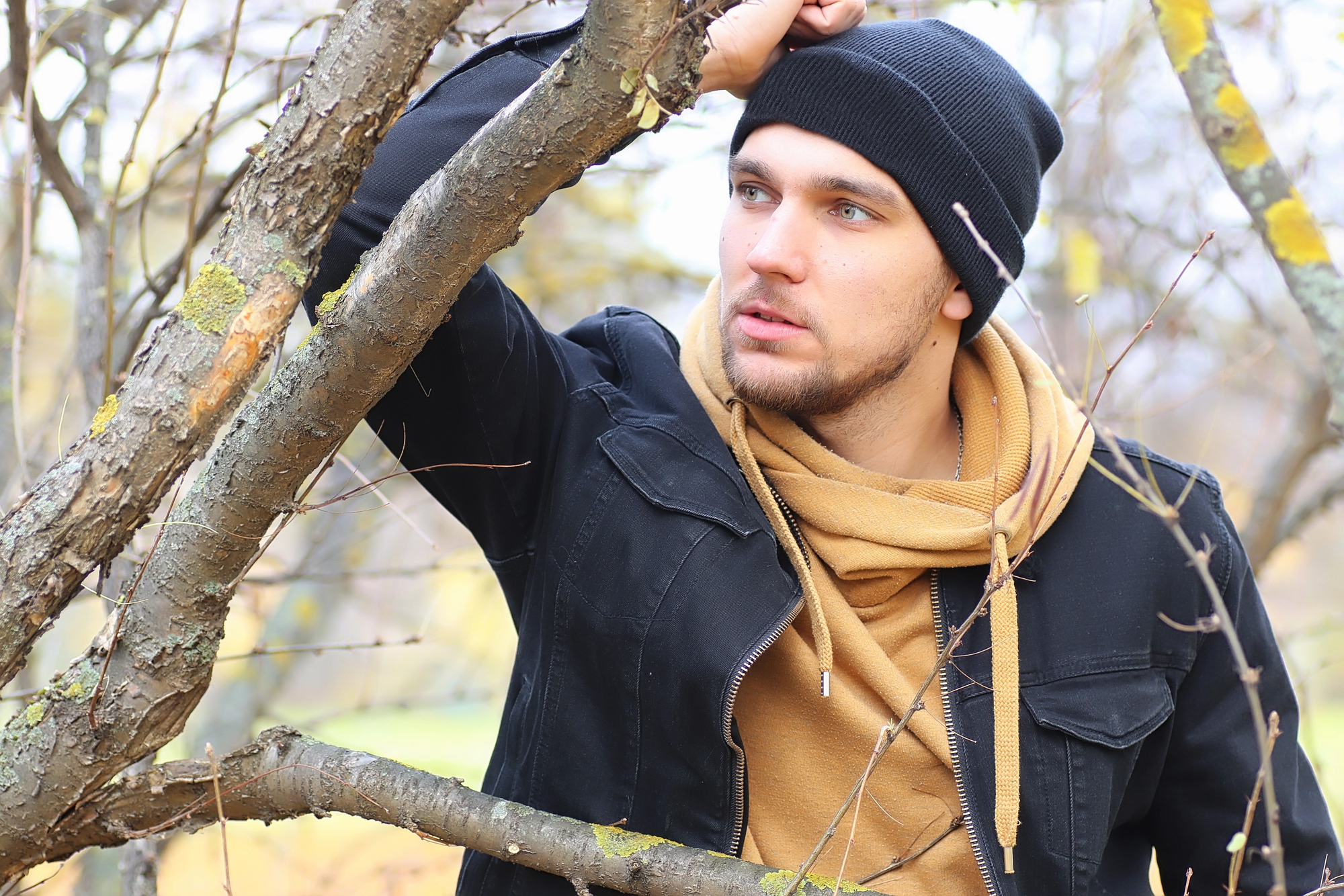 A man wearing a black beanie, a black jacket, and a brown hoodie stands outdoors, leaning on a tree branch with moss. He gazes thoughtfully into the distance. It's a fall day with bare tree branches and light foliage in the background.