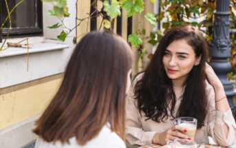 Two women sit at an outdoor café table. One woman with long dark hair holds a glass of coffee, looking towards the other woman, who has light brown hair. They are surrounded by greenery and a building with yellow accents.