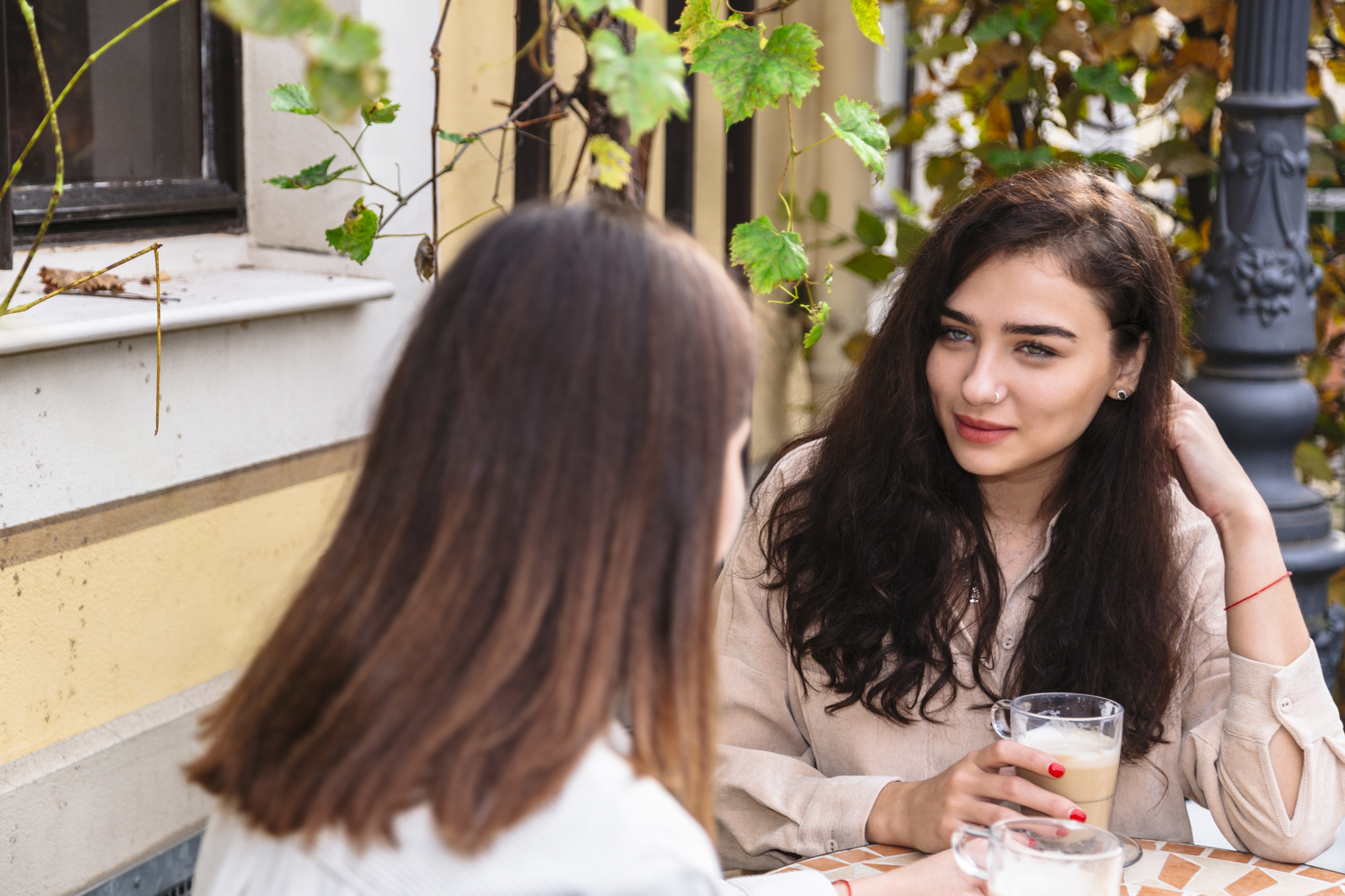 Two women sit at an outdoor café table. One woman with long dark hair holds a glass of coffee, looking towards the other woman, who has light brown hair. They are surrounded by greenery and a building with yellow accents.