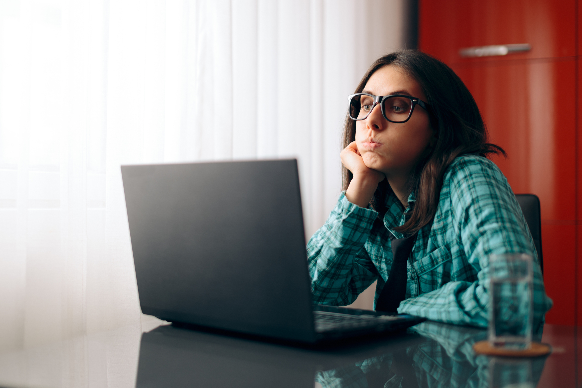 A person wearing glasses and a plaid shirt sits at a desk, looking at a laptop. They rest their chin on their hand, appearing thoughtful. A glass of water is nearby, and a bright window curtain is in the background.