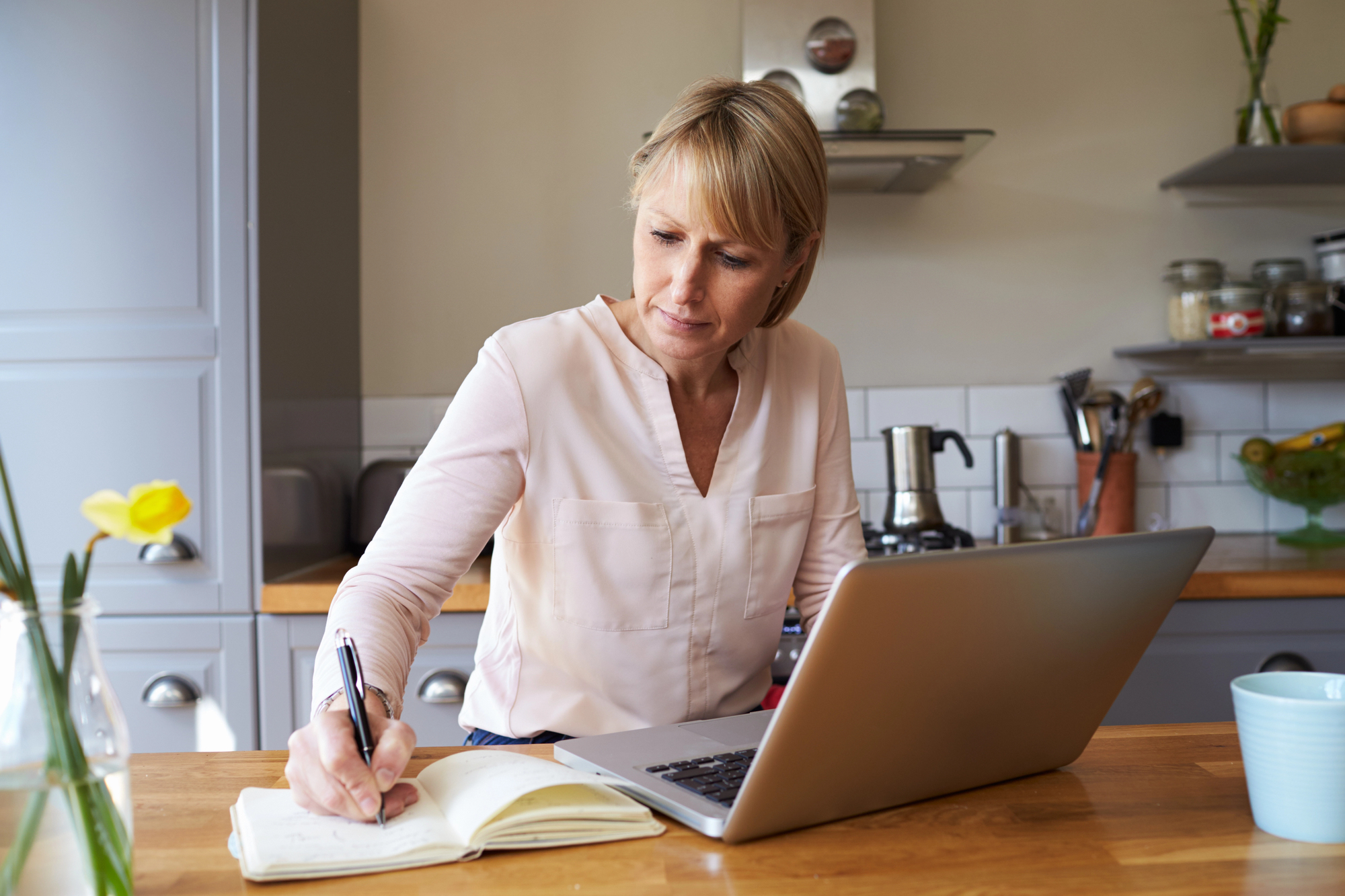 A woman sits at a kitchen table with a laptop, writing in a notebook. The room has gray cabinets and various kitchen items. A yellow flower is in a vase on the table. She appears focused and thoughtful.