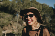 A woman with blonde hair smiles broadly, wearing a wide-brimmed hat, sunglasses, and a black mesh top. She stands outdoors with a backdrop of blurred trees and sunlight filtering through, creating a warm, cheerful atmosphere.