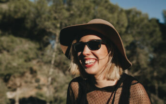 A woman with blonde hair smiles broadly, wearing a wide-brimmed hat, sunglasses, and a black mesh top. She stands outdoors with a backdrop of blurred trees and sunlight filtering through, creating a warm, cheerful atmosphere.