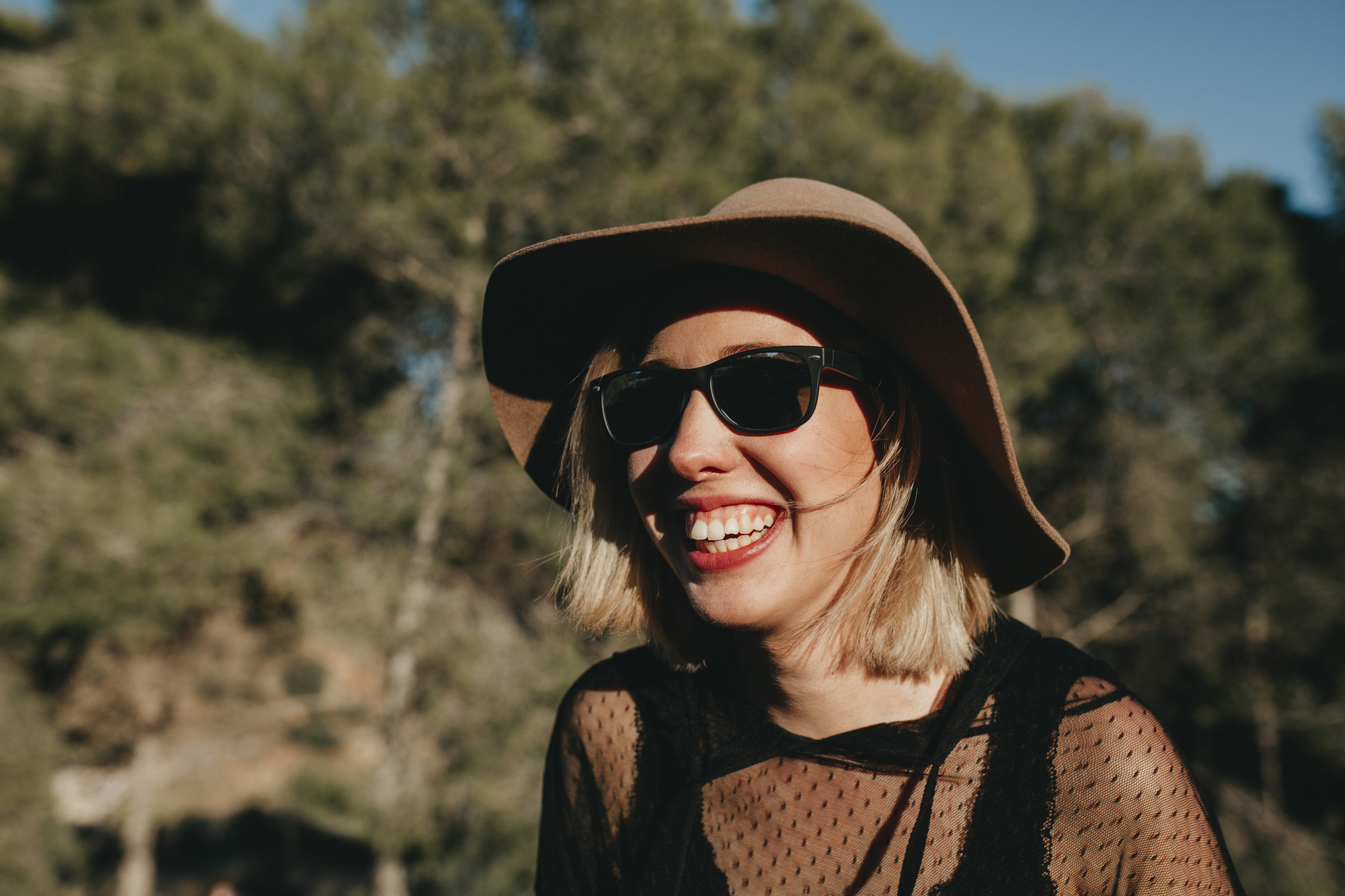 A woman with blonde hair smiles broadly, wearing a wide-brimmed hat, sunglasses, and a black mesh top. She stands outdoors with a backdrop of blurred trees and sunlight filtering through, creating a warm, cheerful atmosphere.