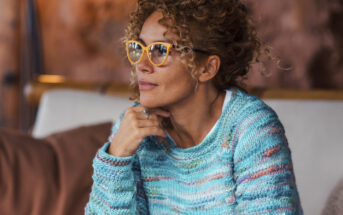 A person with curly hair and glasses is sitting indoors, wearing a colorful knit sweater. They appear thoughtful, resting their chin on their hand, with a blurred background.