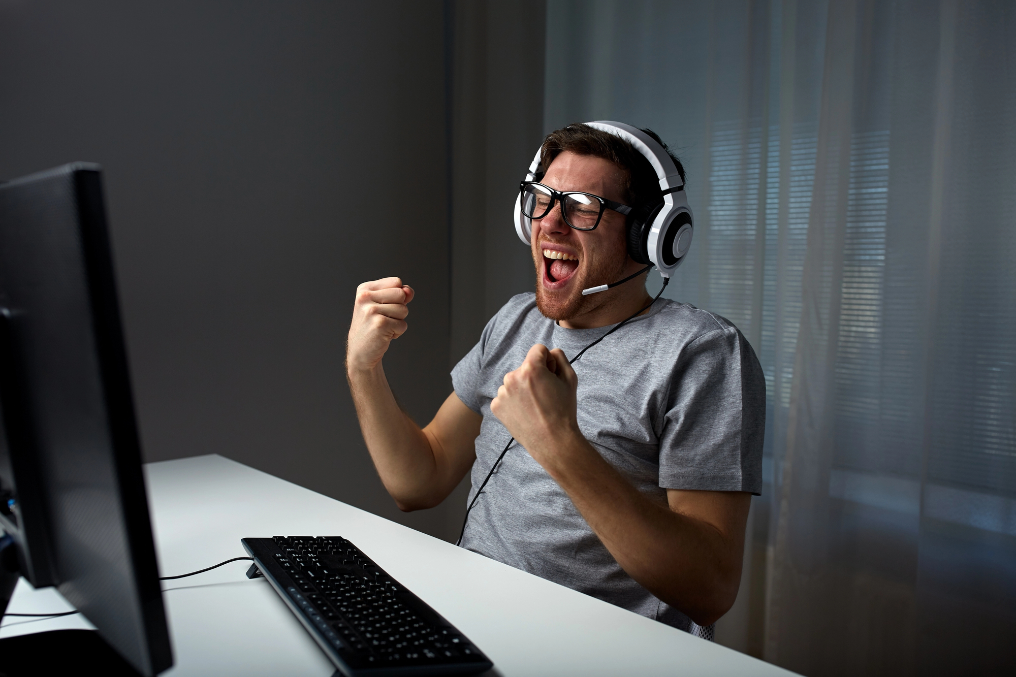 A person wearing glasses and a headset is sitting at a desk in front of a computer, clenching their fists and smiling widely, appearing excited or triumphant. A keyboard and mouse are on the desk.