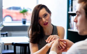 A woman with long brown hair and a checkered top holds hands with a man across a table in a brightly lit cafe. She gazes at him intently, with an orange car visible through the window in the background.