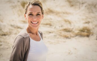 A woman with a smile, wearing a white shirt and beige cardigan, stands on a sunny beach with sand and dried grass in the background.