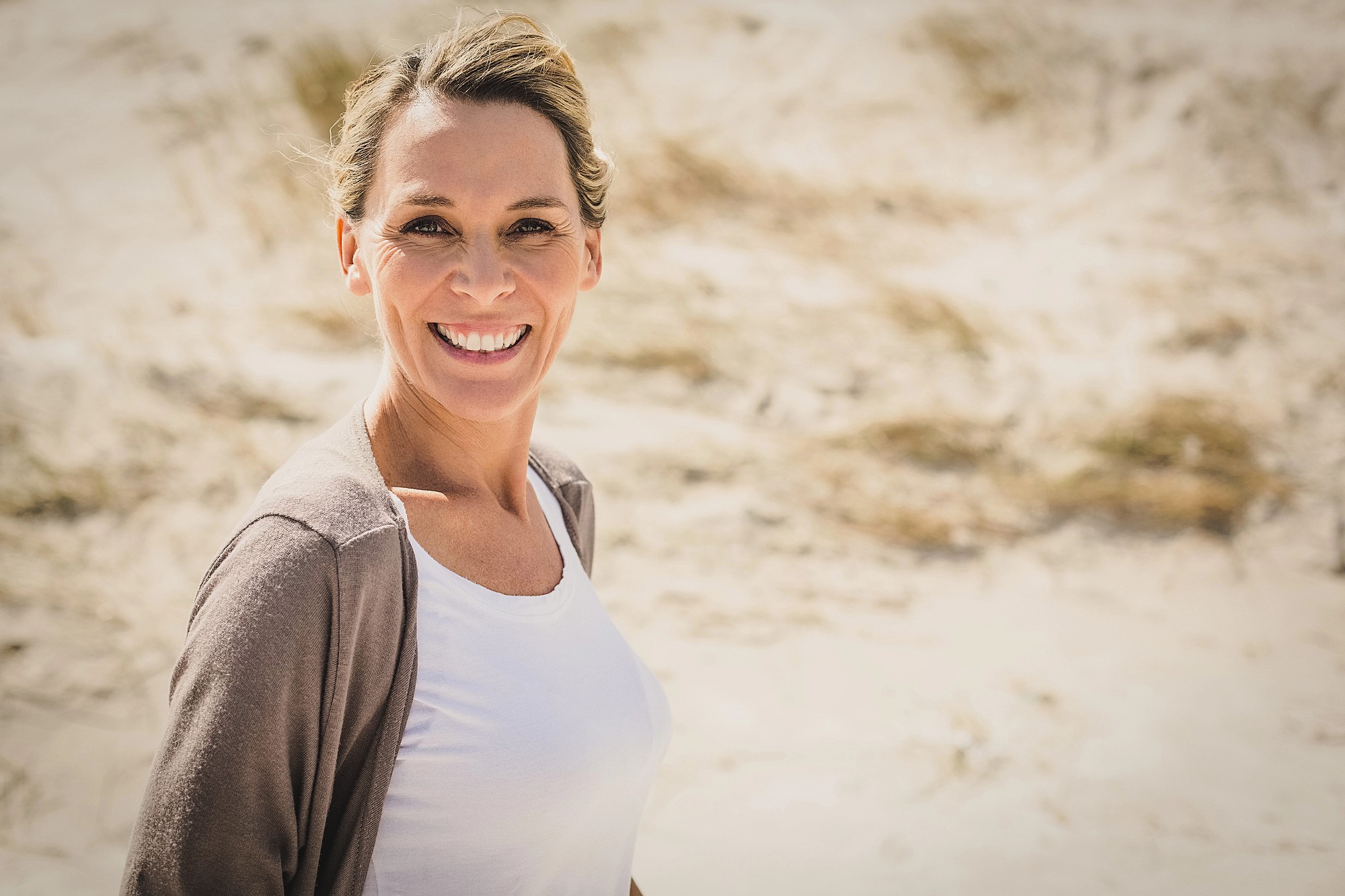 A woman with a smile, wearing a white shirt and beige cardigan, stands on a sunny beach with sand and dried grass in the background.