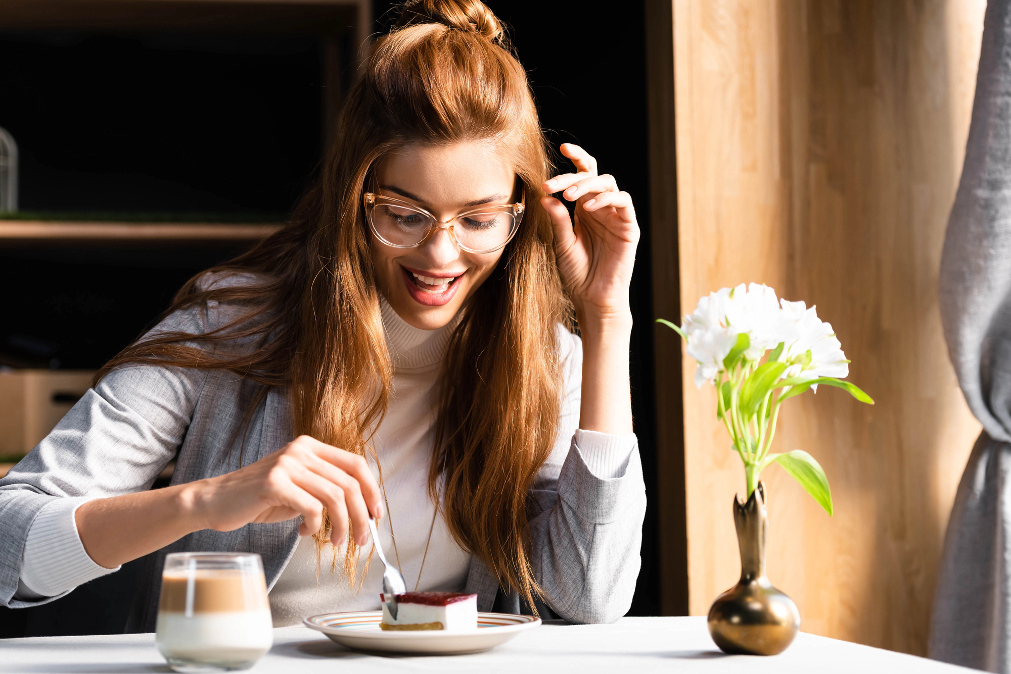 A woman with long hair sits at a table, smiling as she enjoys a dessert on a plate. A glass of beverage is nearby, along with a small vase holding white flowers. Sunlight streams in, creating a warm and cozy atmosphere.