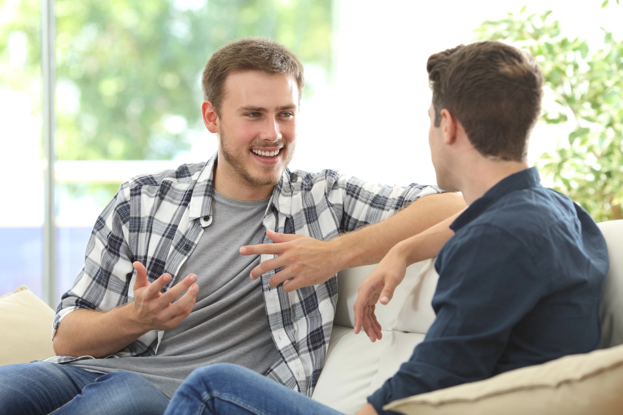 Two men are sitting on a couch in a bright room. One is wearing a plaid shirt and gesturing while talking, smiling at the other man, who wears a dark blue shirt. The background shows blurred greenery and windows, indicating a relaxed setting.