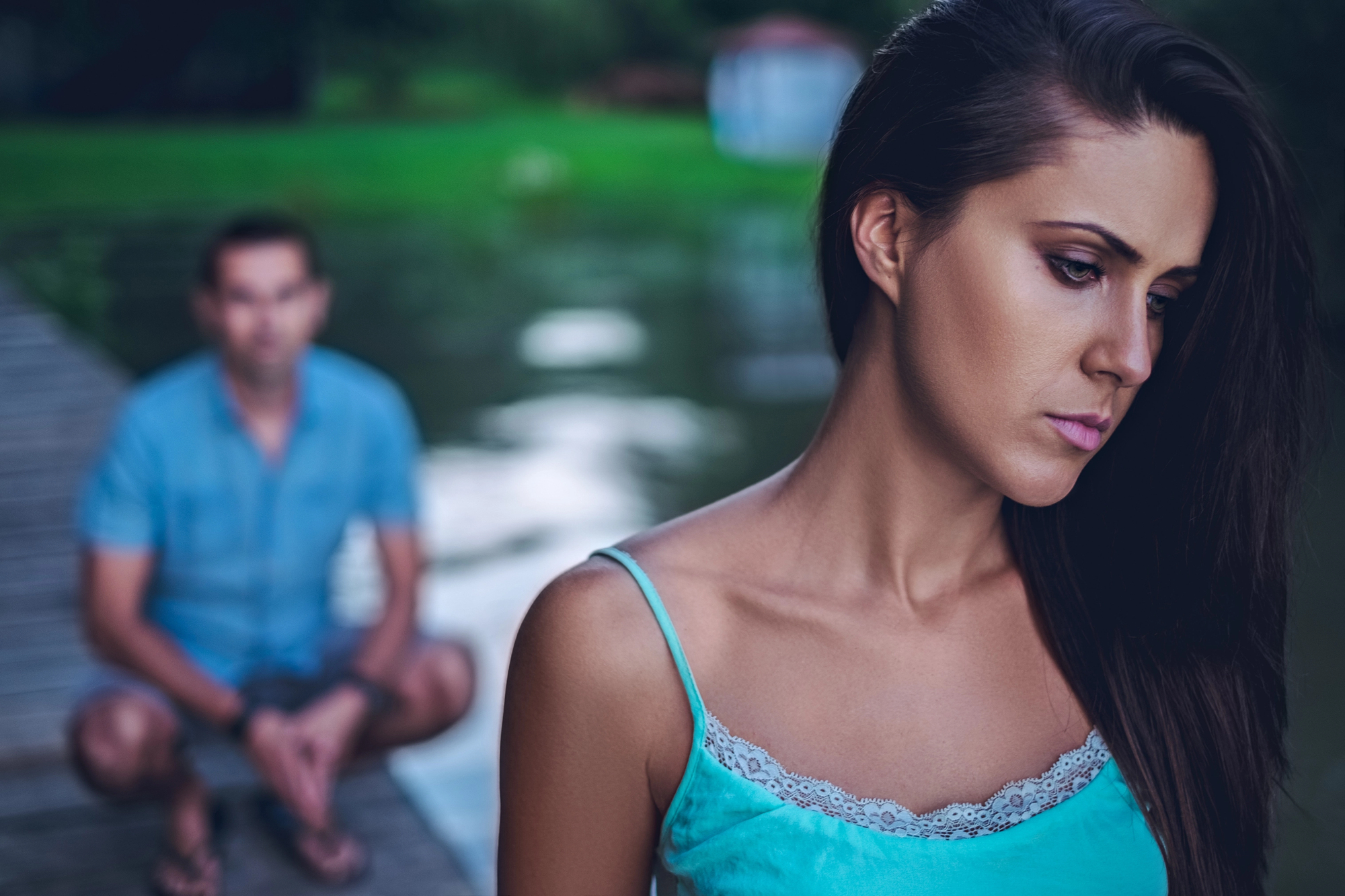 A woman with long brown hair in a blue sleeveless top stands in the foreground, looking pensive and slightly sad. Behind her, a man in a blue shirt and shorts is squatting on a wooden dock near a body of water, out of focus.