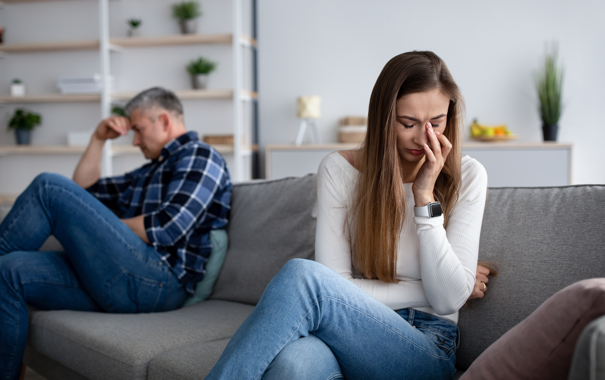 A woman sits on a couch with her hand on her face, appearing upset. A man in the background sits with his head resting in his hand, also looking distressed. The room has neutral colors with shelves and plants.