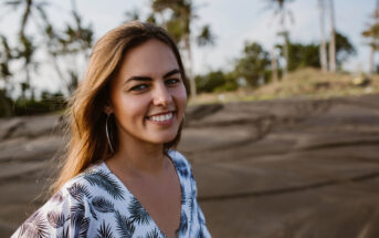 A woman with long hair smiles warmly at the camera. She is wearing a white and black patterned dress. In the background, there are palm trees and a sandy landscape under a clear sky.