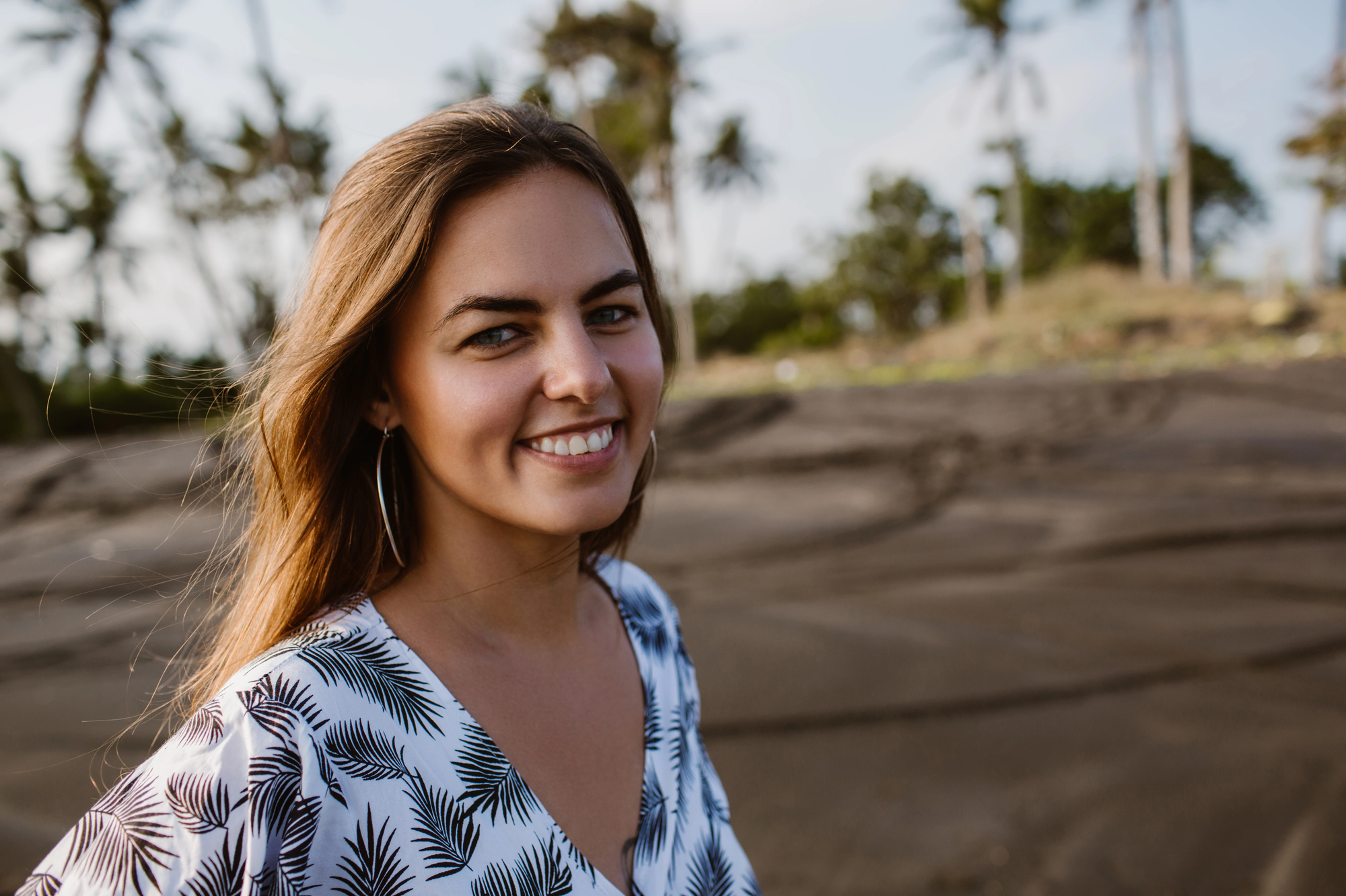 A woman with long hair smiles warmly at the camera. She is wearing a white and black patterned dress. In the background, there are palm trees and a sandy landscape under a clear sky.