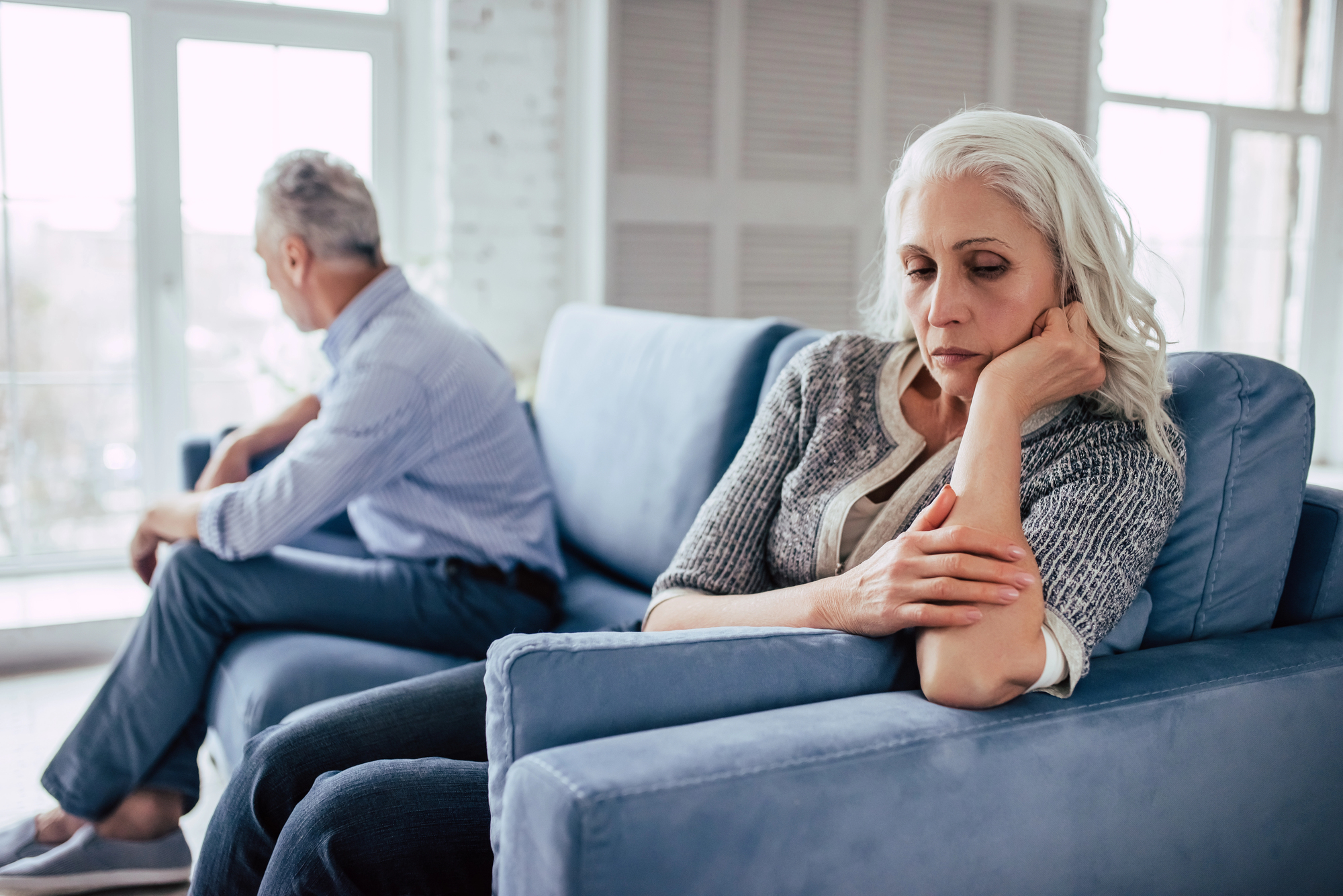 An older couple sits on a blue couch in a bright room, both appearing upset. The woman in the foreground is looking down, with her hand on her cheek. The man in the background is turned away, looking out the window.