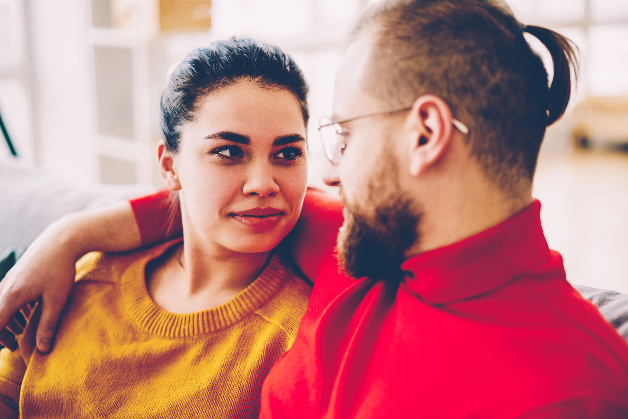 A couple sits close together on a couch, gazing into each other's eyes with gentle smiles. The woman has dark hair tied back and wears a yellow sweater. The man with a beard and glasses wears a red sweater, and his arm rests around the woman's shoulders.