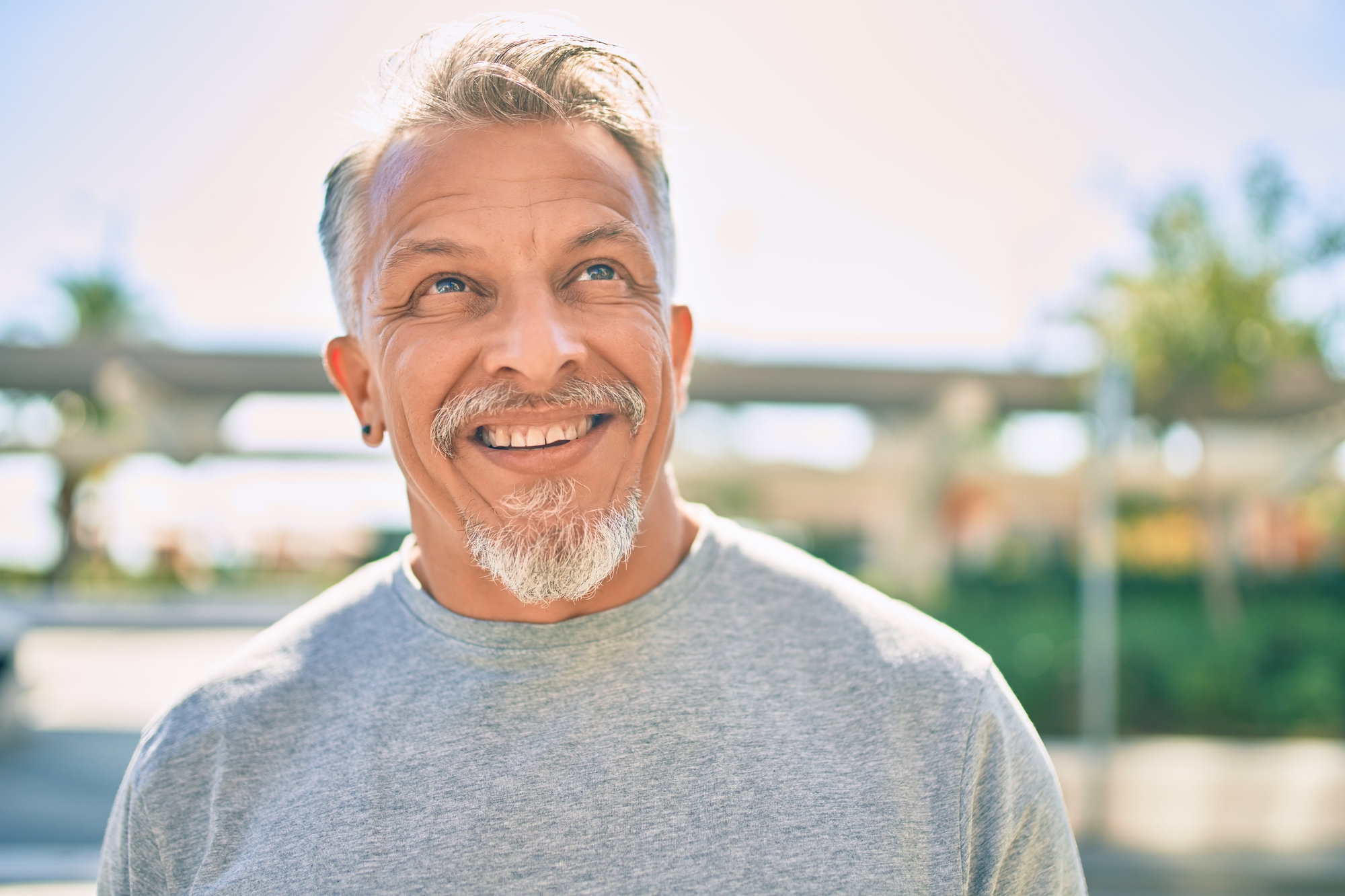 A middle-aged man with gray hair and a beard smiles while looking up. He is wearing a gray T-shirt and standing outdoors, with a blurred background of greenery and structures on a sunny day.