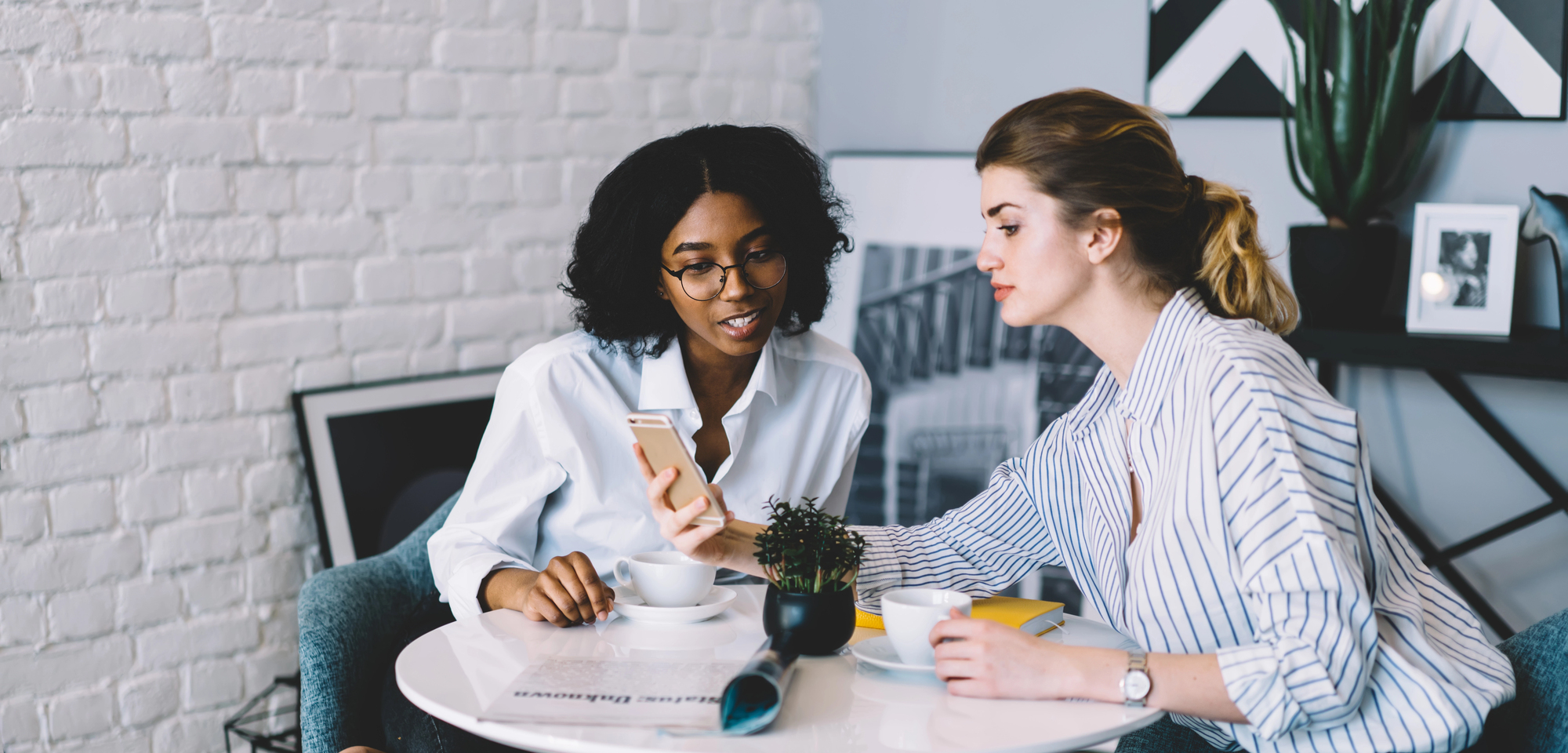 Two women sitting at a round table, engaged in conversation. One holds a smartphone, showing it to the other. They each have a cup of coffee in front of them, with a small potted plant on the table. Cozy room with art on the walls.