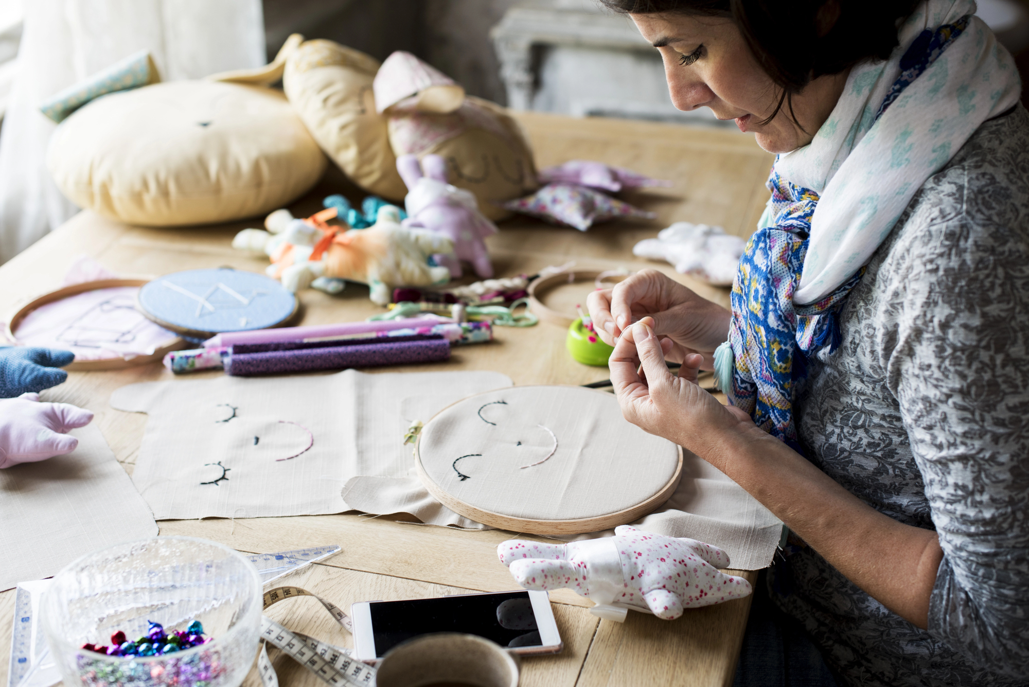 A woman is sitting at a table embroidering a smiling face on fabric inside a hoop. The table is covered with sewing materials, plush toys, and a smartphone. She is wearing a scarf and concentrating on her work.