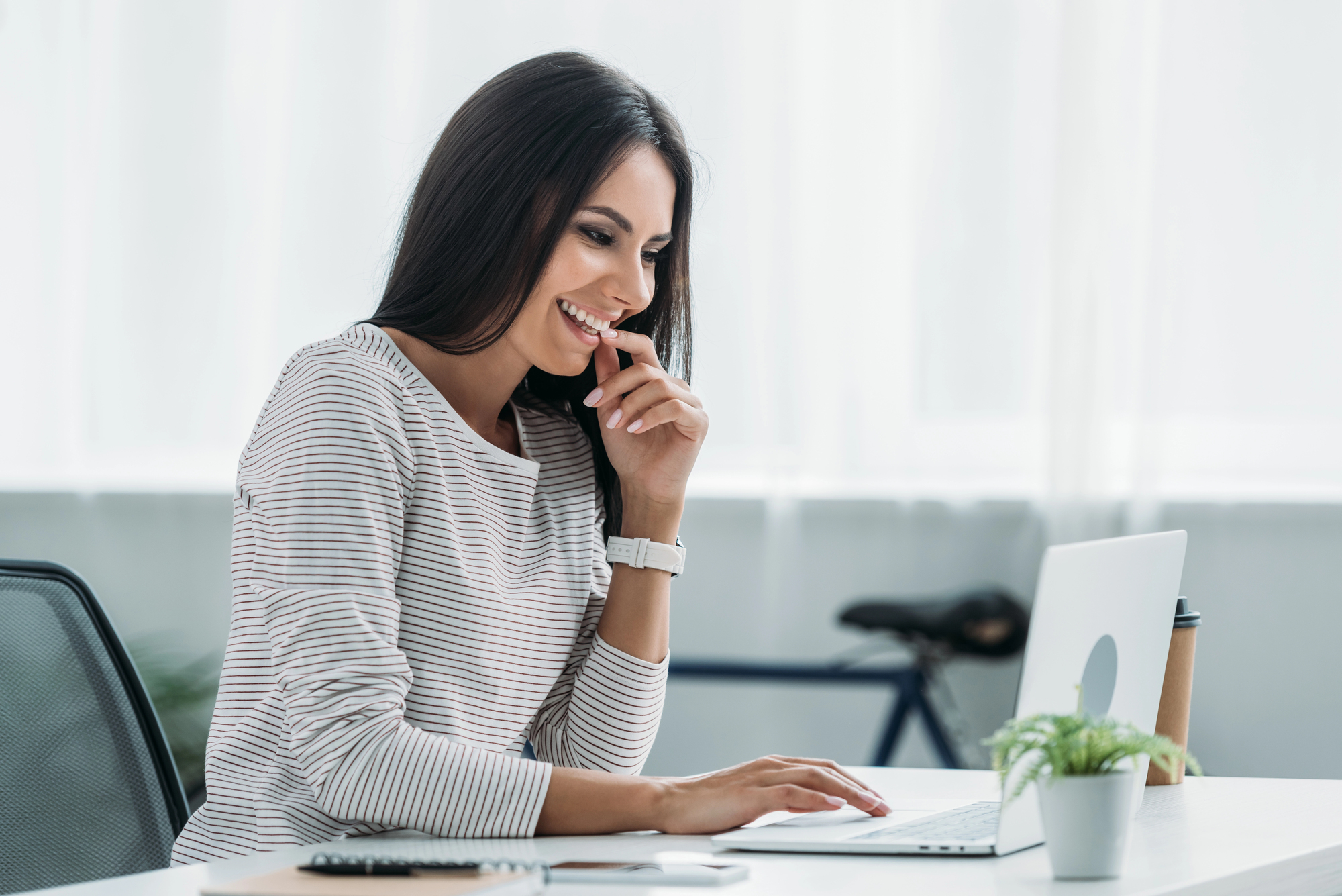 A woman in a striped shirt is sitting at a desk, looking at her laptop with a smile. She is resting her chin on her hand. A plant, keyboard, and cup are on the desk, and a bicycle is in the background.