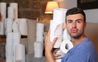 A man in a blue shirt is sitting indoors, holding several rolls of toilet paper close to his face with a playful expression. In the background, there are more rolls stacked on a couch and a lit lamp.