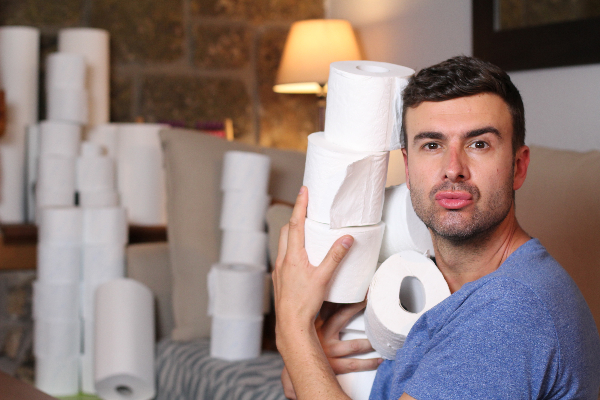 A man in a blue shirt is sitting indoors, holding several rolls of toilet paper close to his face with a playful expression. In the background, there are more rolls stacked on a couch and a lit lamp.