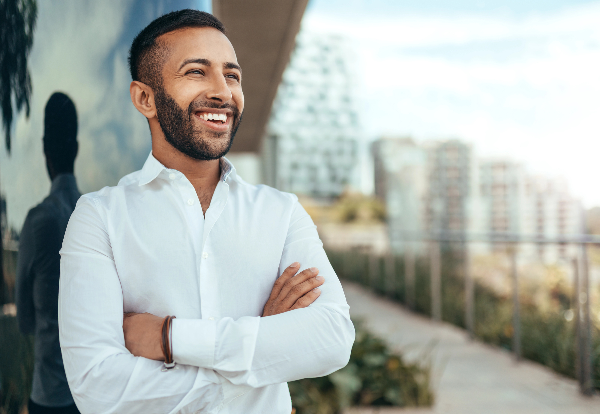 A man in a white shirt stands outdoors with his arms crossed, smiling. The background features a cityscape with modern buildings and a glass wall reflecting part of his image. The atmosphere is bright and sunny.