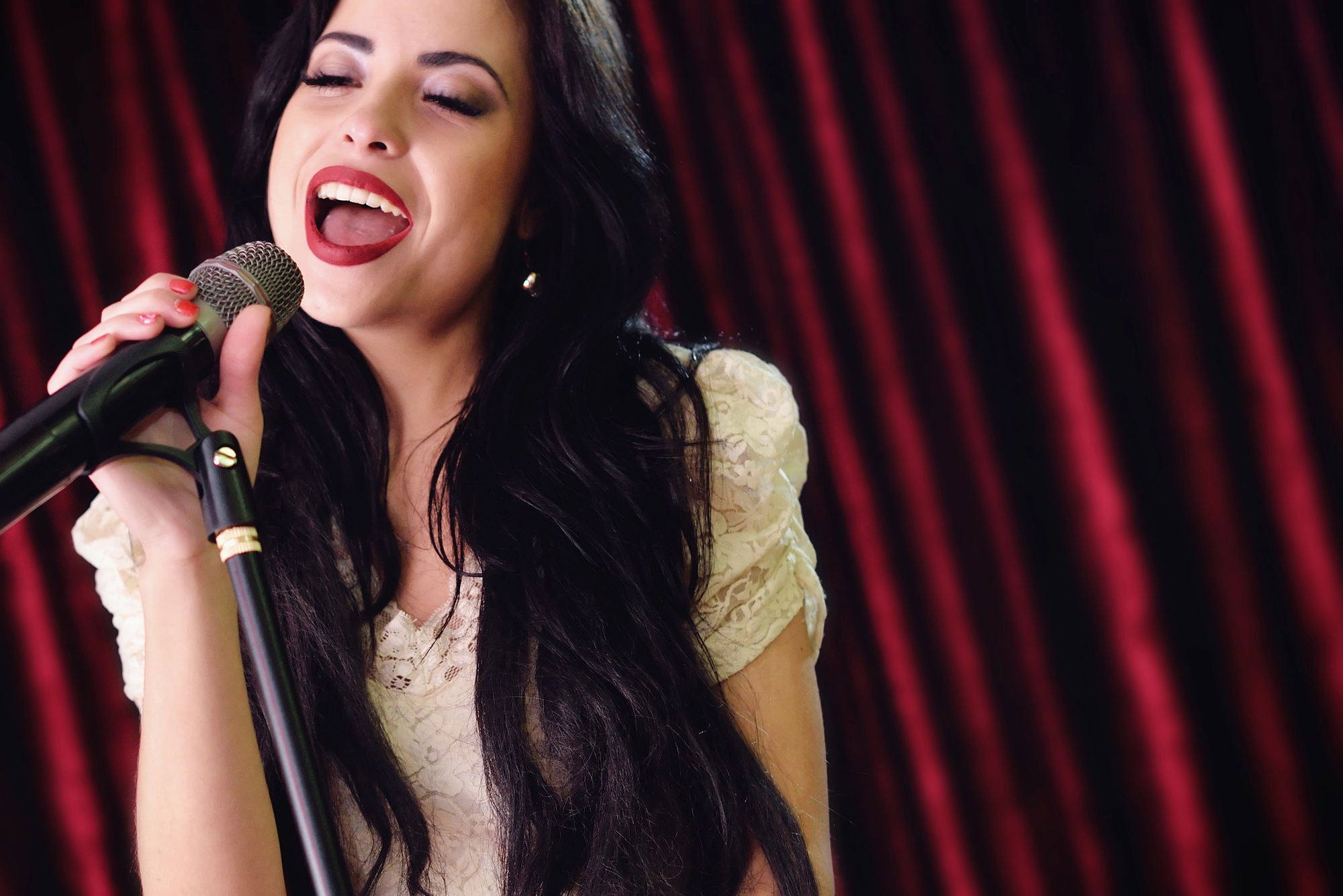 A woman with long dark hair, wearing a white lace top, sings passionately into a microphone. She stands in front of red stage curtains.