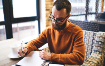 A man with glasses and a beard sits at a table in a cozy cafe, writing in a notebook. He is wearing an orange sweater. A smartphone and a cup of coffee are placed on the table. Natural light filters through large windows.