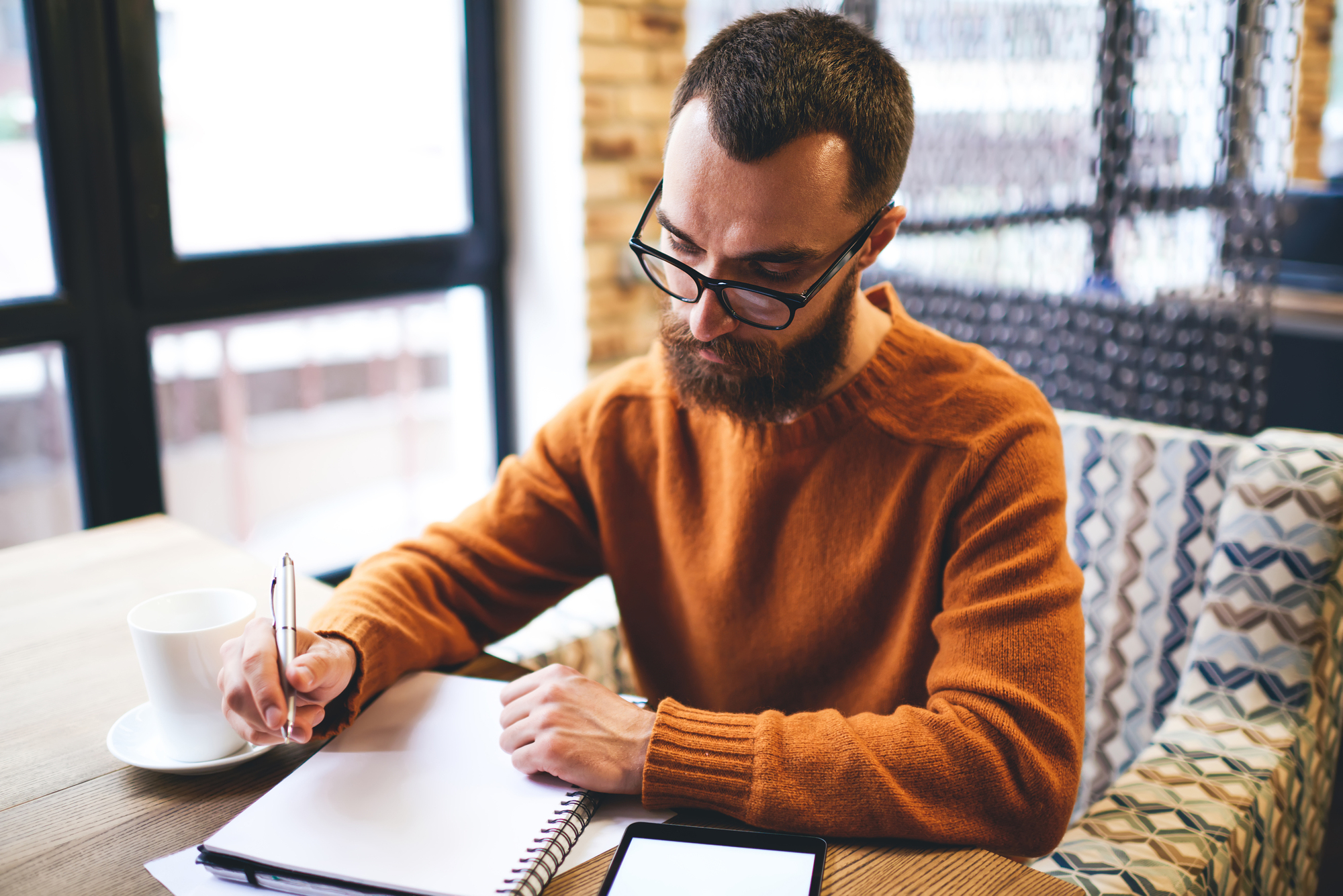 A man with glasses and a beard sits at a table in a cozy cafe, writing in a notebook. He is wearing an orange sweater. A smartphone and a cup of coffee are placed on the table. Natural light filters through large windows.