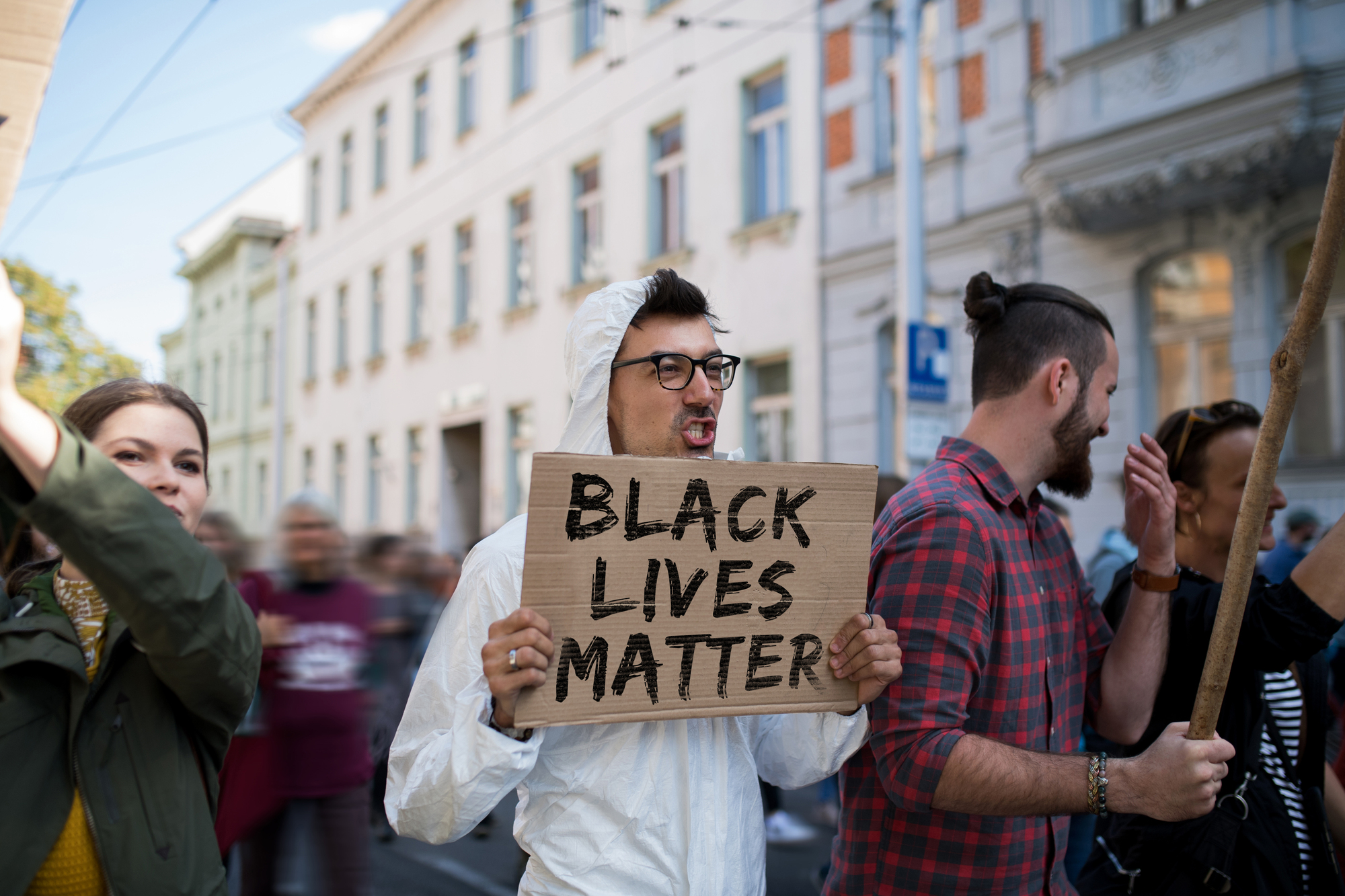 A person holding a "Black Lives Matter" sign stands among a group of protesters. The scene is on a city street with blurred buildings in the background. The person wears glasses and a white hoodie, and people around them are holding up banners.