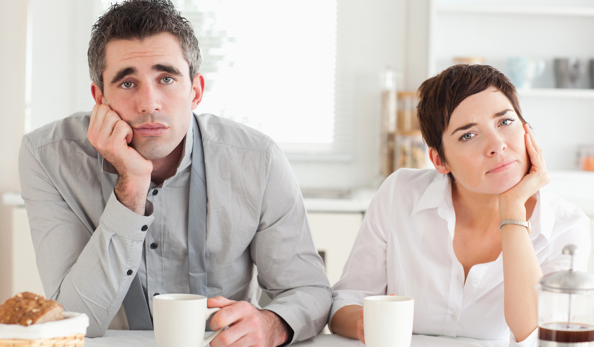 A man and a woman sit at a table, both resting their heads on their hands, appearing bored. They have coffee cups in front of them in a bright kitchen setting.