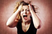 A woman with wavy hair is screaming with her eyes closed, holding her head in her hands against a textured pinkish background, expressing frustration or stress.