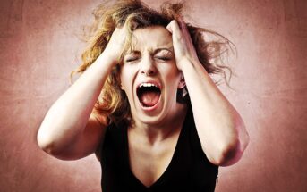 A woman with wavy hair is screaming with her eyes closed, holding her head in her hands against a textured pinkish background, expressing frustration or stress.