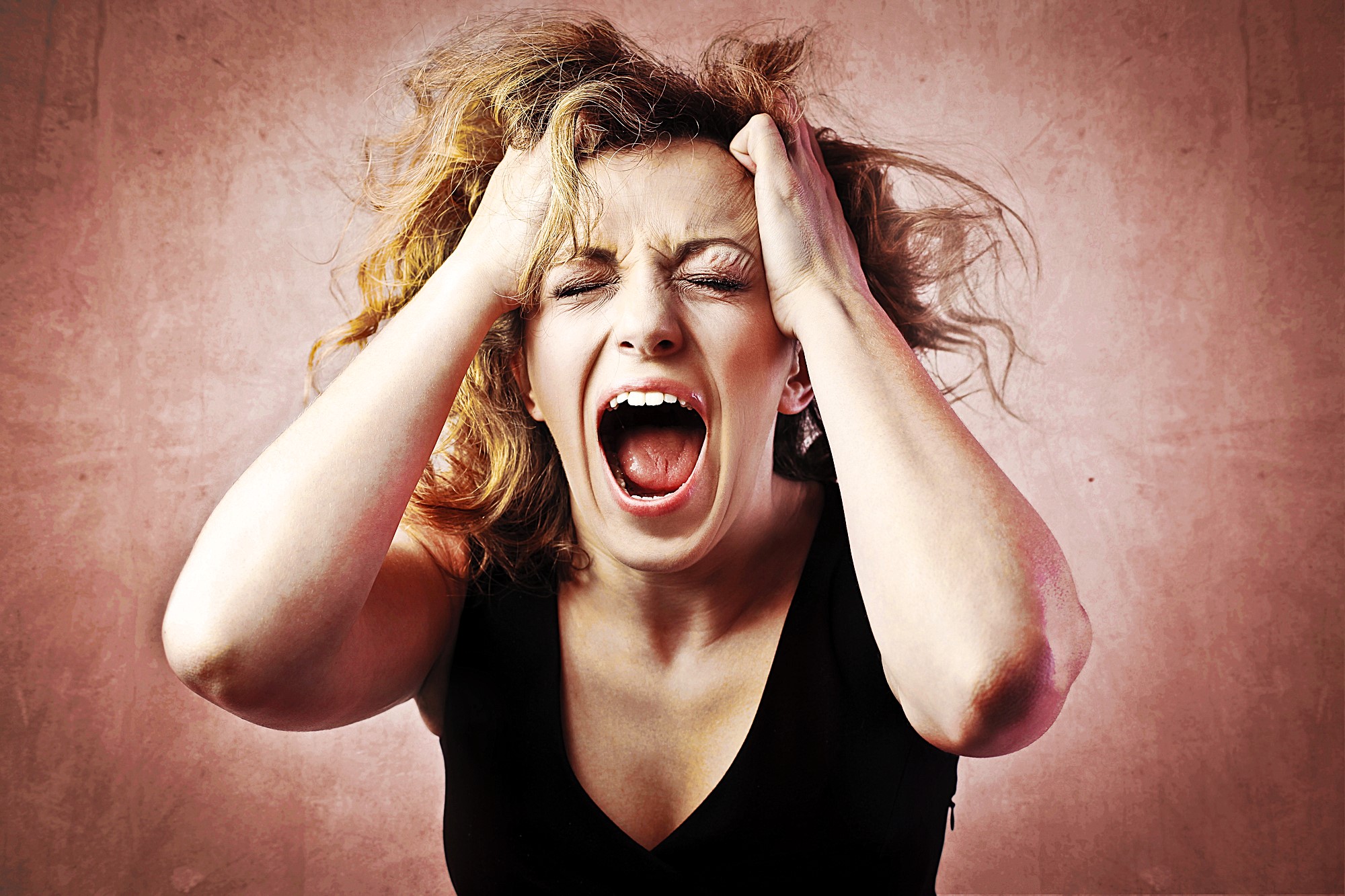 A woman with wavy hair is screaming with her eyes closed, holding her head in her hands against a textured pinkish background, expressing frustration or stress.