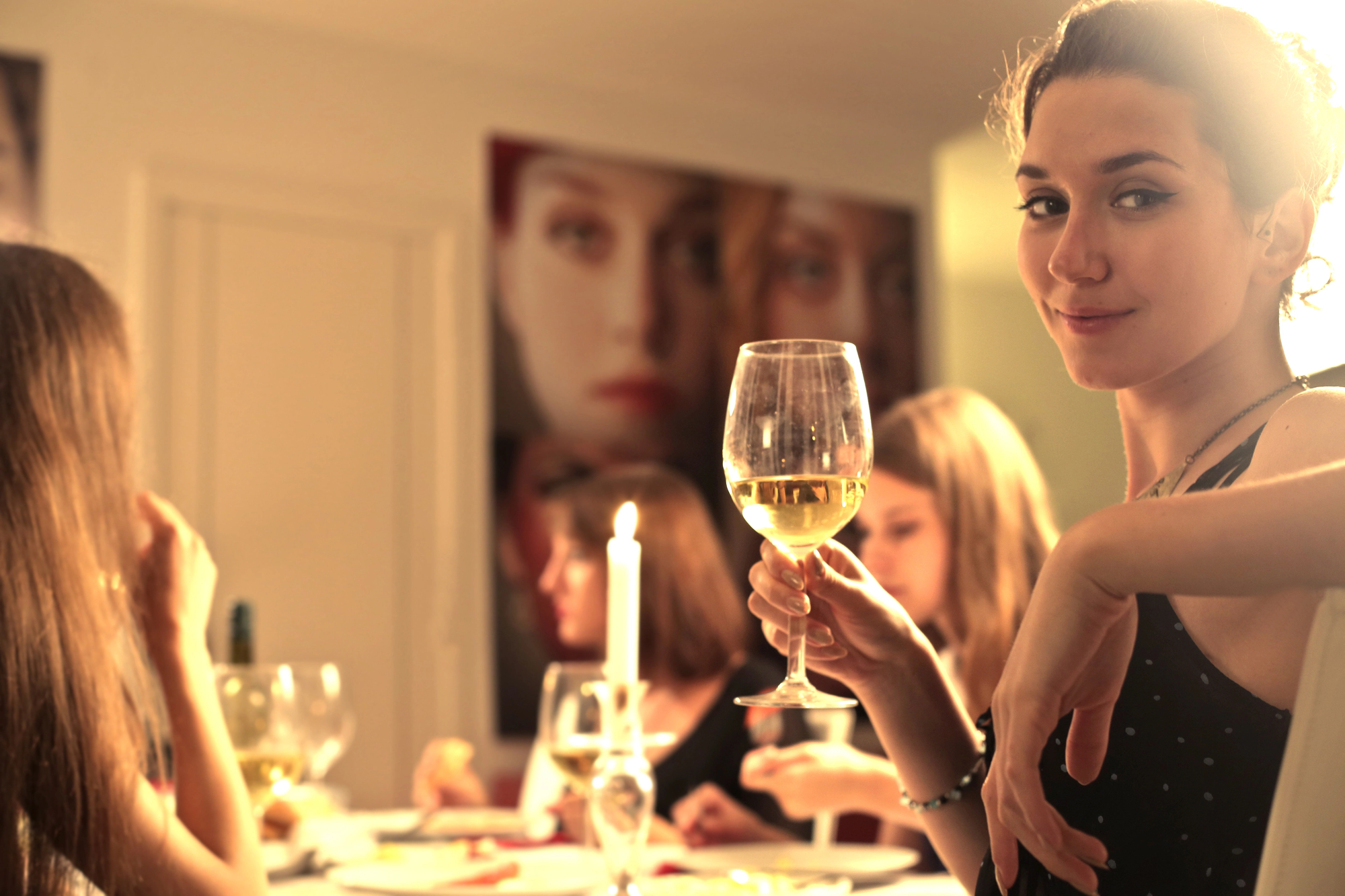 A woman smiles and holds up a wine glass at a dinner table with four other people. The table is set with plates and a lit candle. The background shows blurred images of faces on the wall.