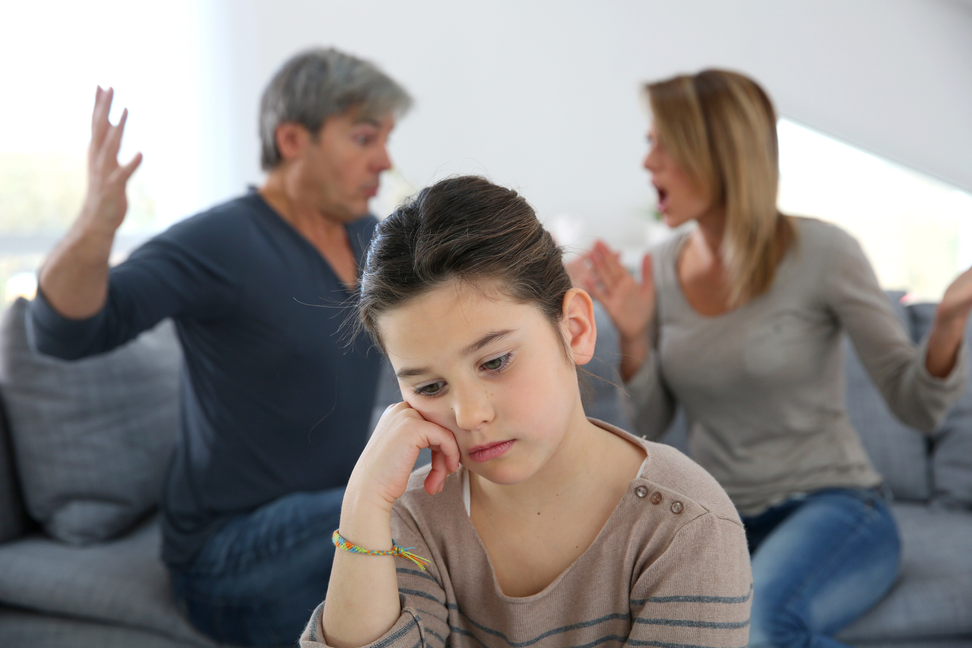 A young girl sits looking sad, resting her chin on her hand, while two adults in the background are arguing energetically. The scene seems tense, highlighting the girl's emotional distress.