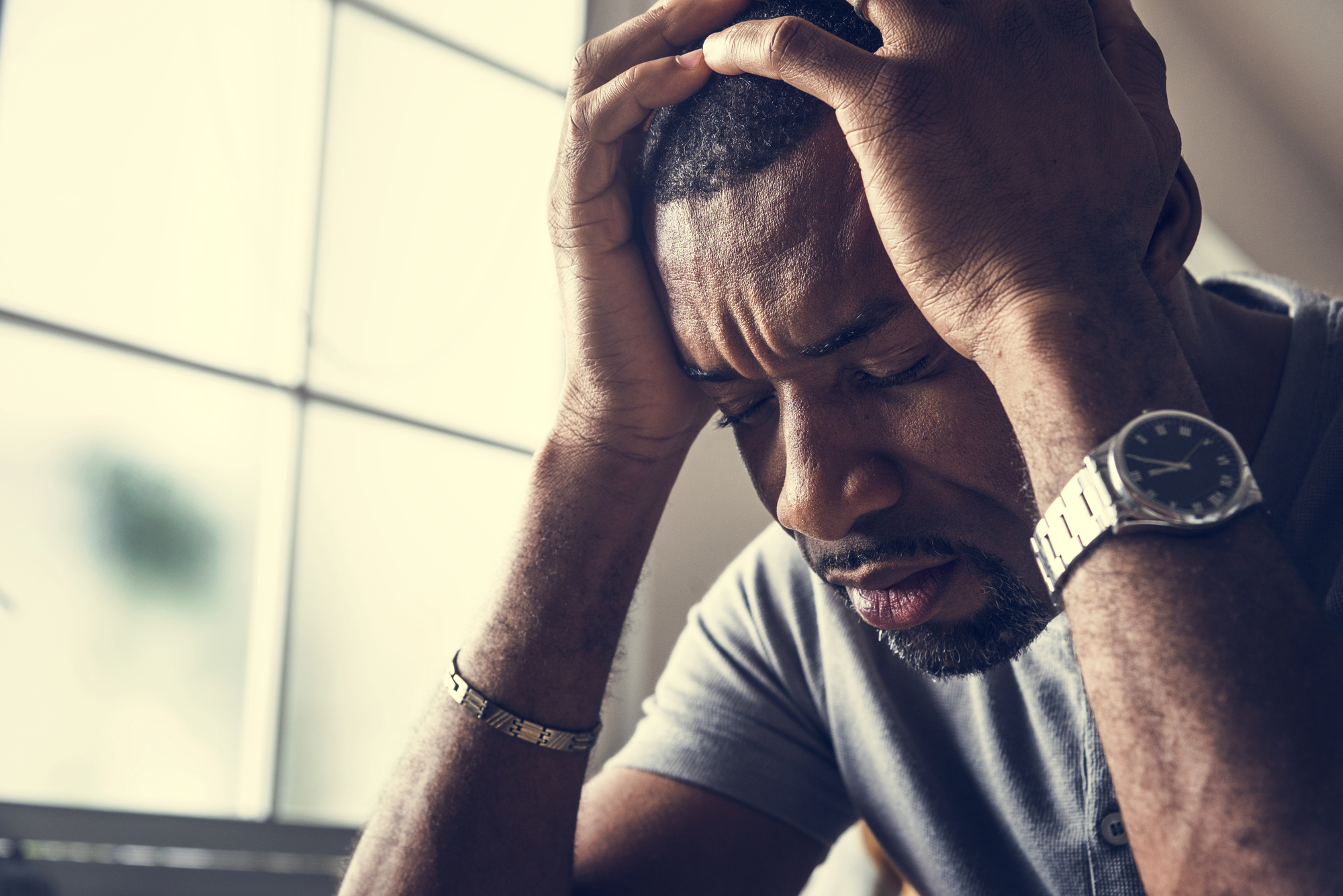 A man in a gray shirt sits indoors with his head in his hands, appearing stressed or frustrated. He wears a watch and bracelet. Sunlight filters through a window in the background.