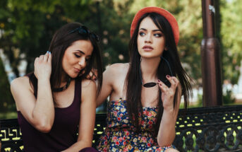 Two women sitting on a bench outdoors. One wears a burgundy dress, adjusting her hair, the other in a floral dress and orange hat, holding sunglasses. They are surrounded by greenery, creating a relaxed and summery atmosphere.