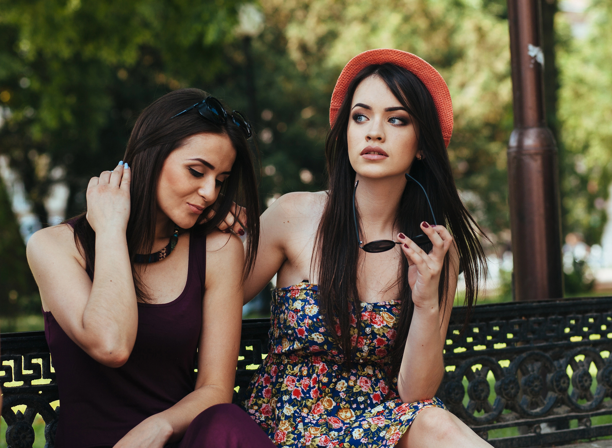 Two women sitting on a bench outdoors. One wears a burgundy dress, adjusting her hair, the other in a floral dress and orange hat, holding sunglasses. They are surrounded by greenery, creating a relaxed and summery atmosphere.