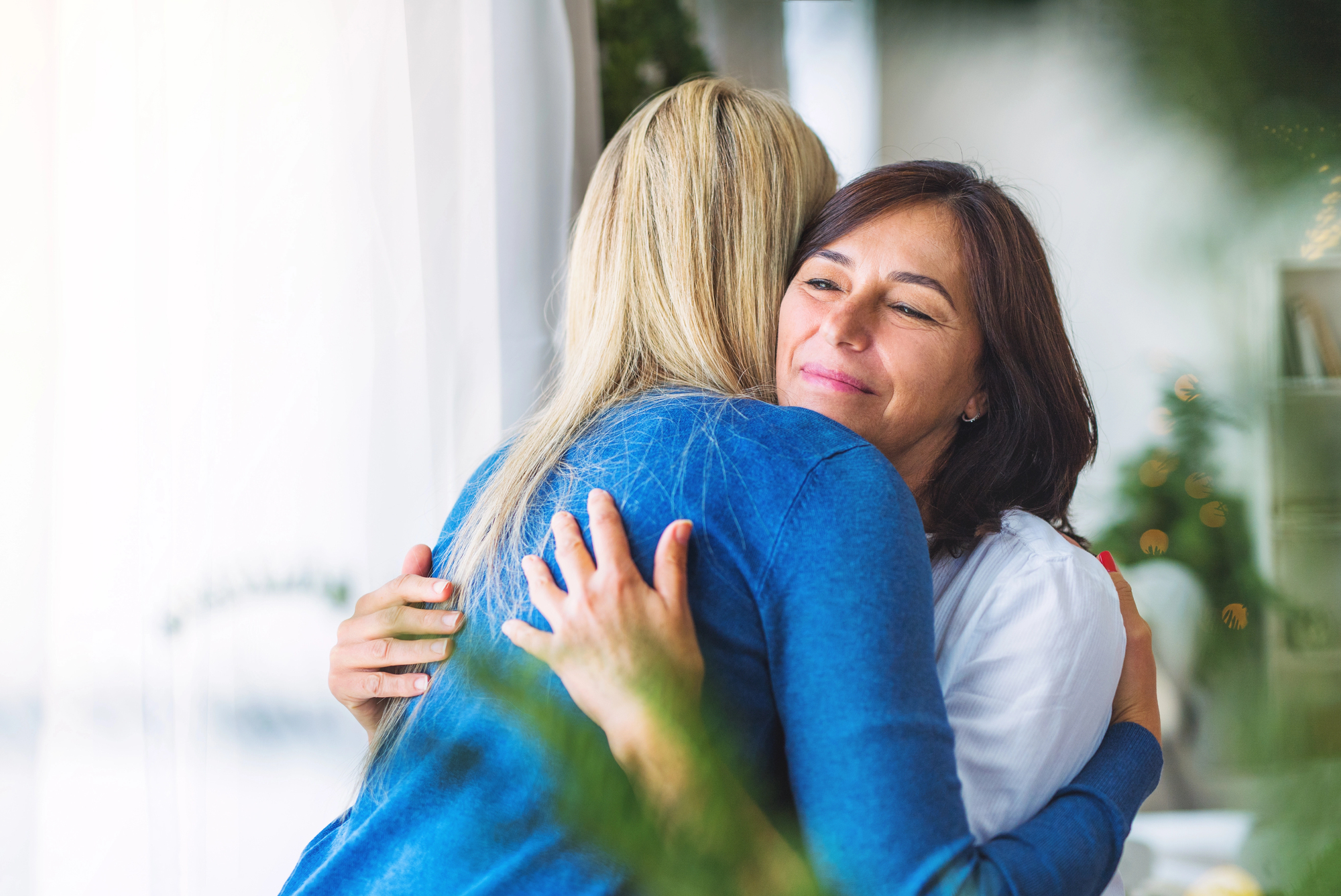 Two women are hugging warmly indoors. One woman, with long blonde hair, is facing away, and the other woman, with short brown hair, is smiling with her eyes closed. They appear to be sharing a comforting or joyful moment.