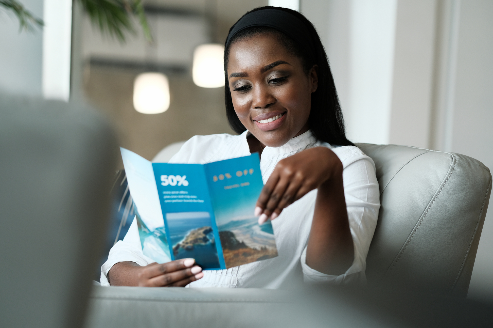 A woman sitting on a couch smiles while reading a travel brochure. She is wearing a white blouse and a black headband. The brochure features beach imagery and a 50% discount offer. A blurred background with lights is visible.