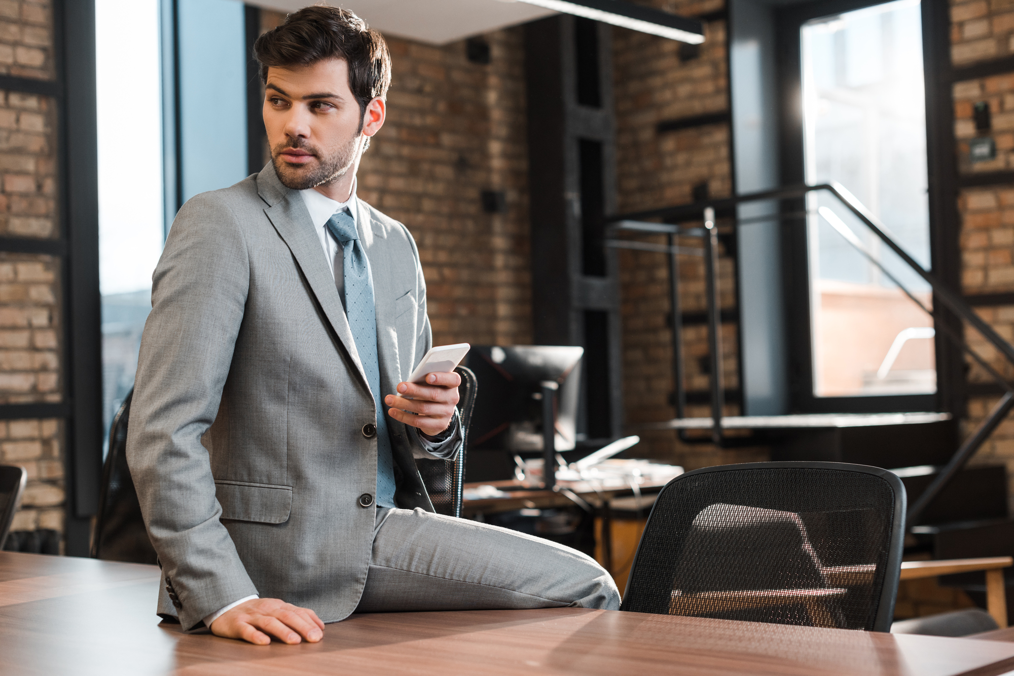 A man in a gray suit sits on a desk in a modern office space holding a smartphone. He is looking to the side with a thoughtful expression. The background features brick walls and large windows.