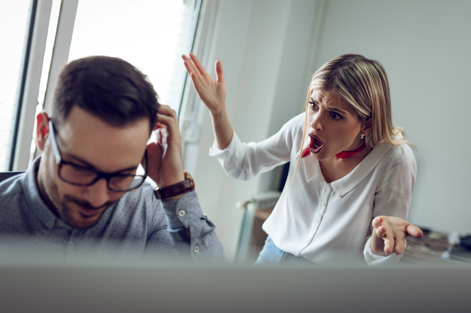 A woman is gesturing and speaking loudly, appearing upset, while a man sits at a desk in front of a computer, holding his head with one hand and looking down. They are in an office setting with a bright window behind them.