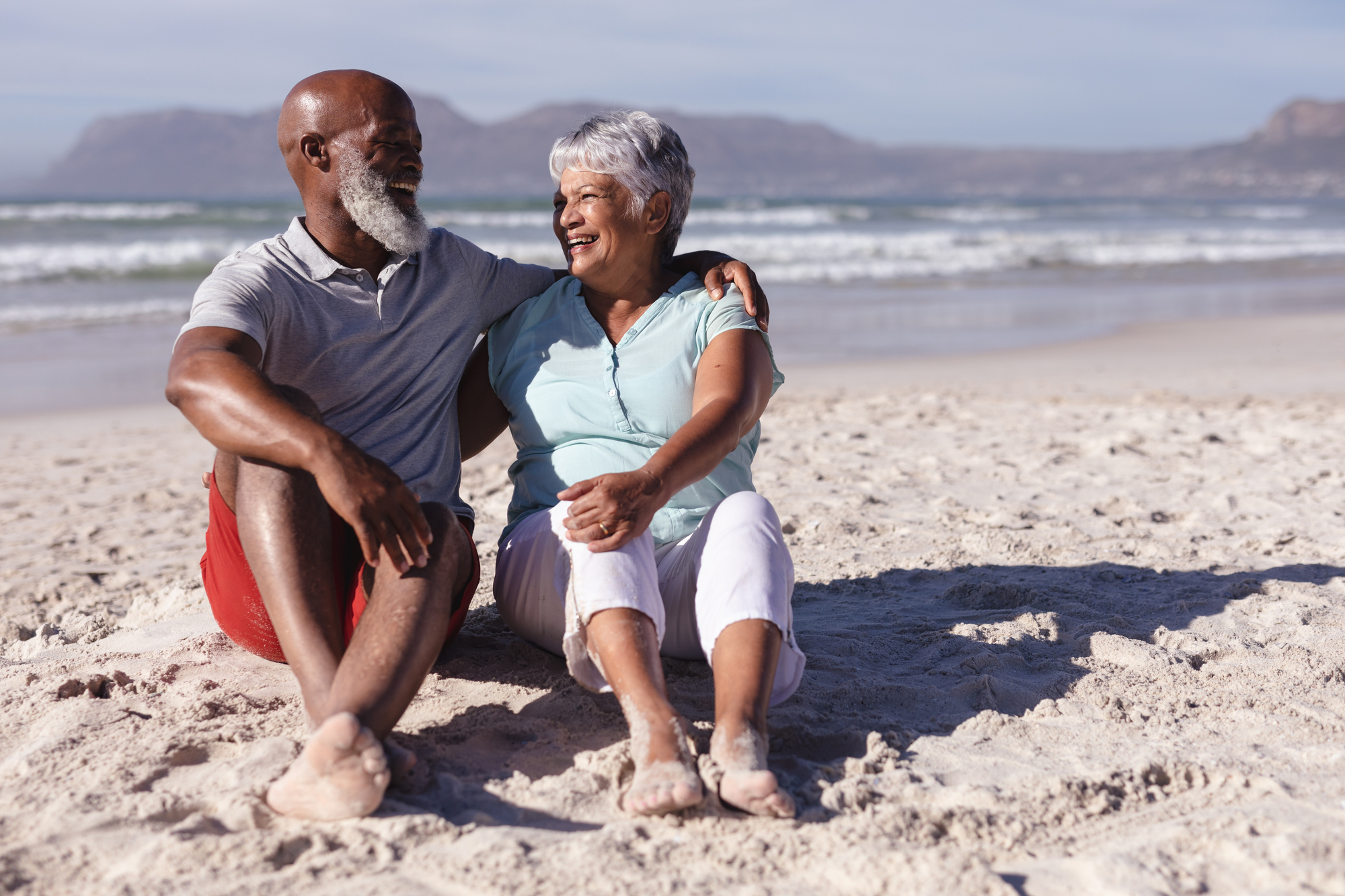 A smiling elderly couple sits on a sandy beach with their arms around each other. They are looking at each other, enjoying the sunny day, with the ocean and distant mountains in the background.