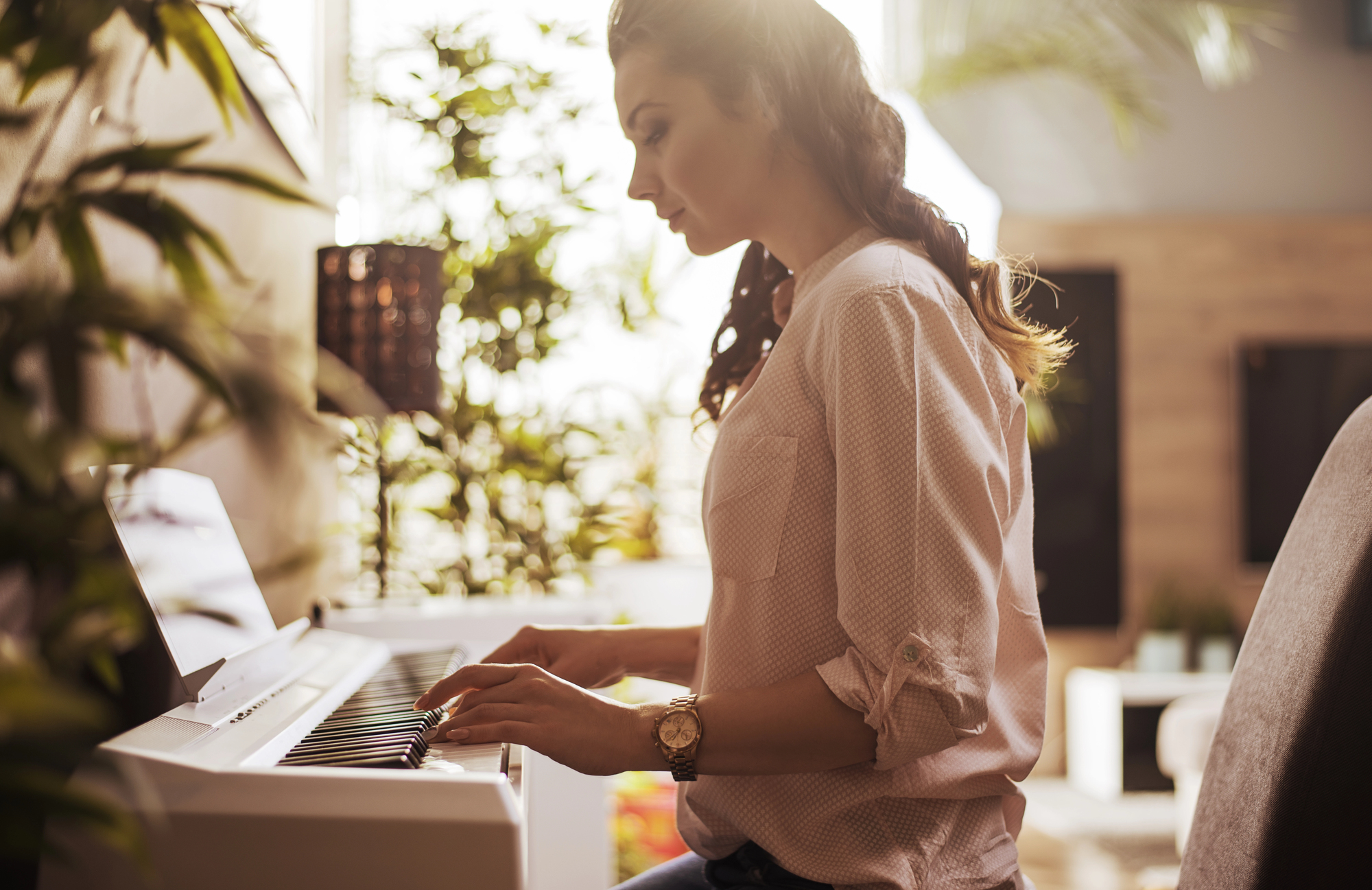 A woman plays an electric piano in a cozy, sunlit room with plants and soft furnishings. She wears a light blouse and a watch, with her hair tied back.