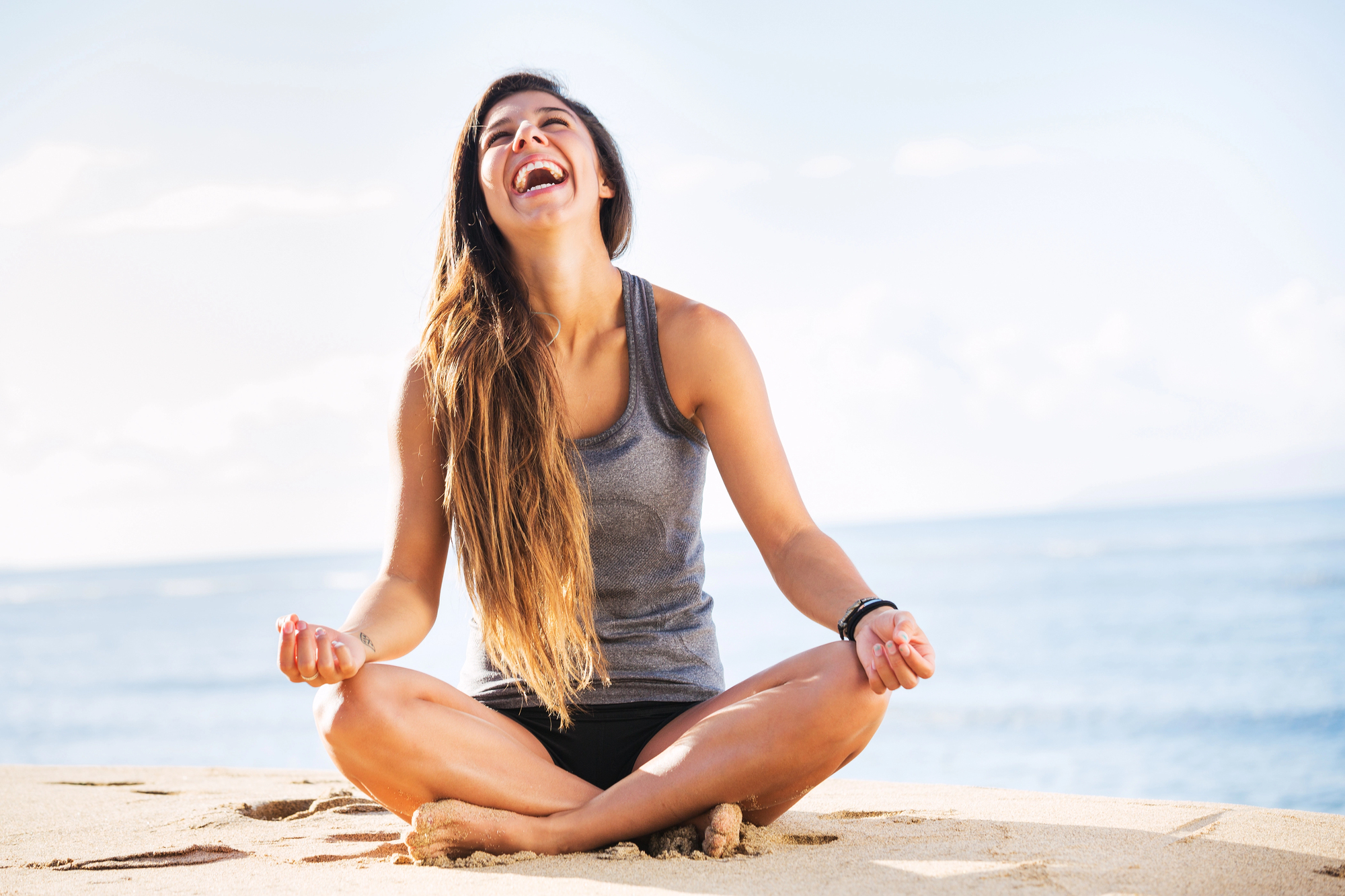 A woman sitting cross-legged on a sandy beach, laughing joyfully with eyes closed. She is wearing a gray tank top and black shorts. The ocean and a clear sky are in the background.