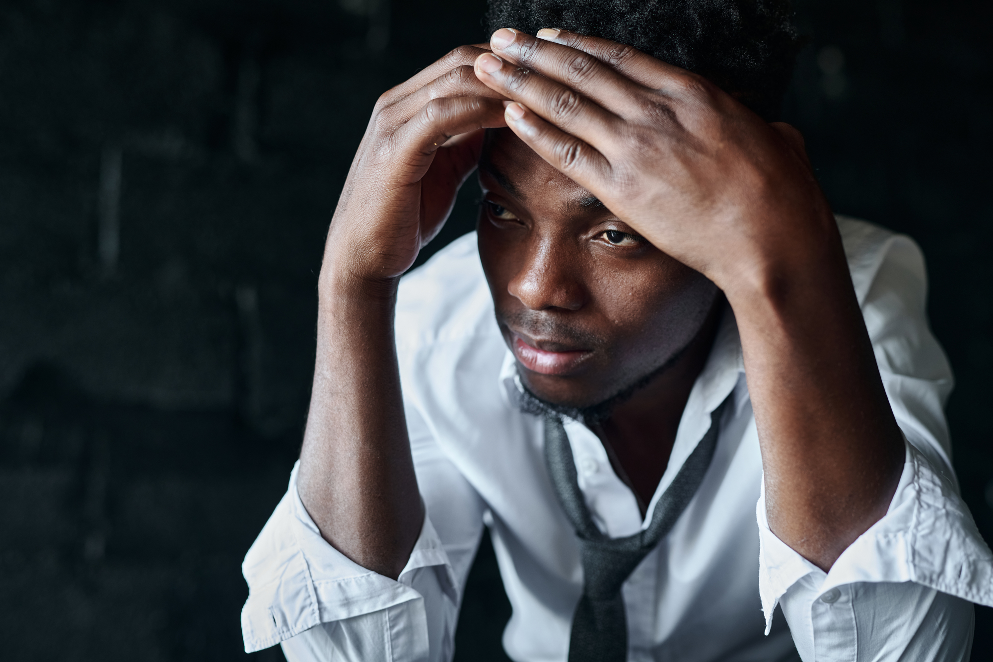 A man in a white dress shirt and loosened tie leans forward, resting his head in his hands. He gazes ahead with a thoughtful expression against a dark background.